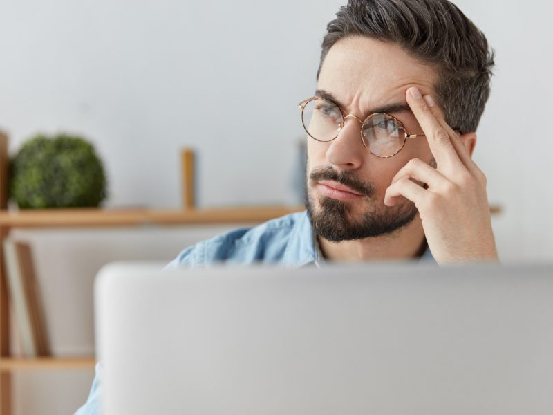Horizontal shot of bearded stylish male copywriter thinks about content of new article, sits in fron Horizontal shot of bearded stylish male copywriter thinks about content of new article, sits in fron